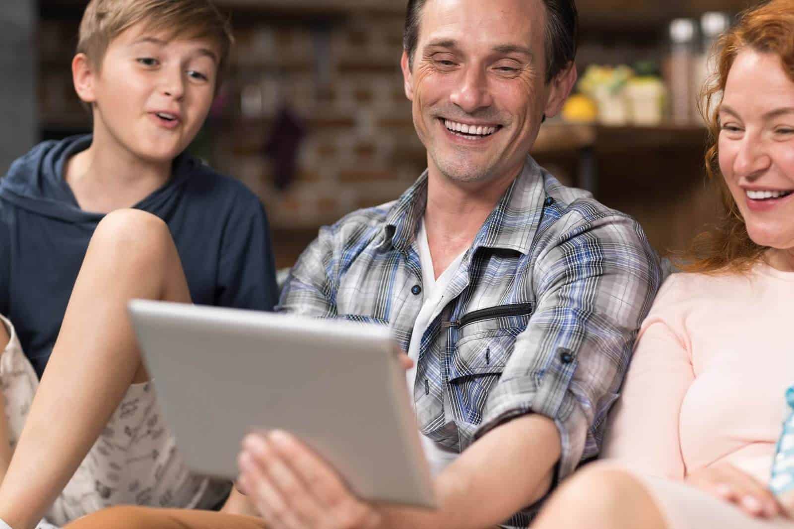 Family Use Tablet Computer Sitting On Couch In Living Room, Parents Spending Time With Son